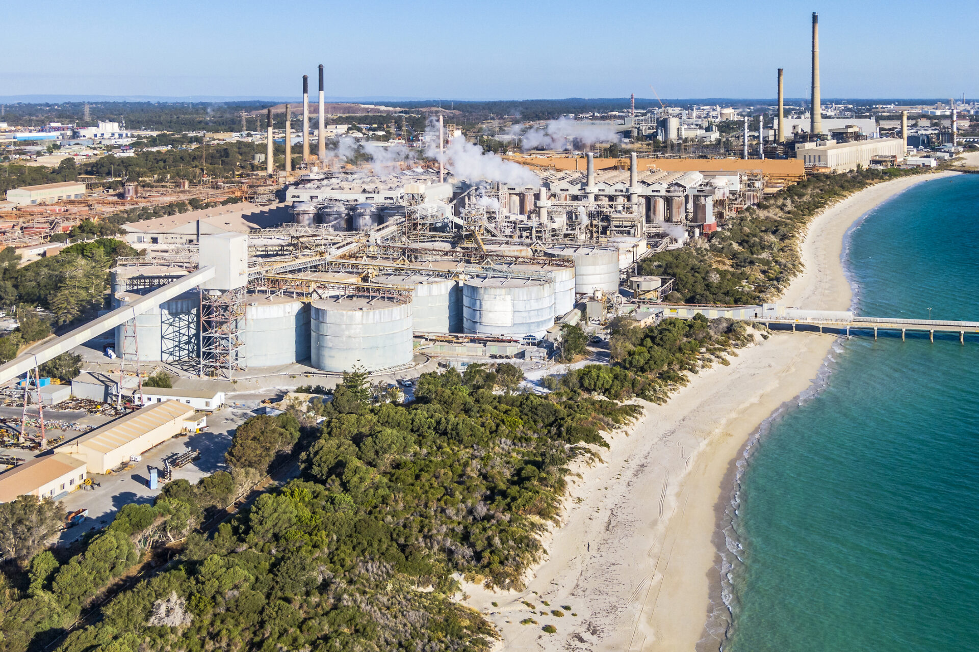 Aerial view alumina refinery in close proximity to natural coastal scrub, clean sandy beach and clear turquoise Indian Ocean sea. Large manufacturing complex with jetty jutting out to sea.