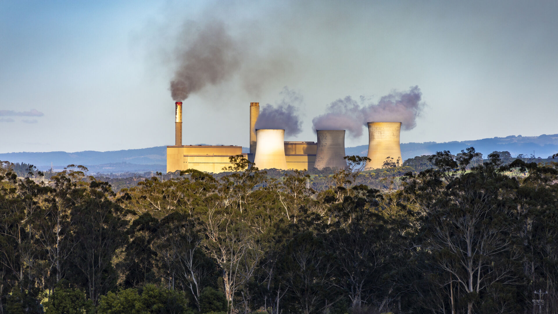 Brown coal-fired power station amongst native trees and hills, emitting smoke from twin smokestacks and heat from three cooling towers: Yallourn Power Station in Latrobe Valley, Victoria to be closed in 2028 and be replaced by a huge battery: climate change, global warming, harmful to health, environmental issues.